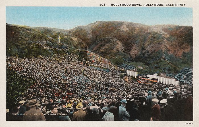 A historic image capturing a bustling event at the Hollywood Bowl, situated among hills, with a large crowd facing a stage set for an upcoming performance.