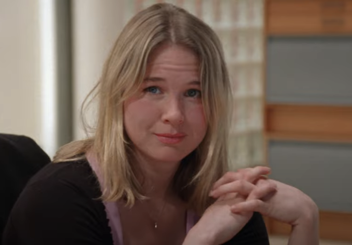 Renee as Bridget, with long hair, sitting indoors, wearing a dark top and gazing thoughtfully at the camera