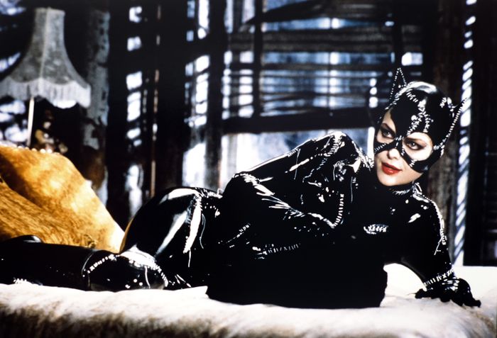 A person is lounging on a bed indoors, wearing a shiny, form-fitting cat-themed costume with a mask and ears
