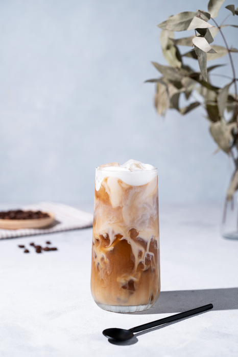 Iced coffee latte served in a tall glass with milk, accompanied by coffee beans, a spoon, and morning shadows. A perfect summer refresher. Front view.