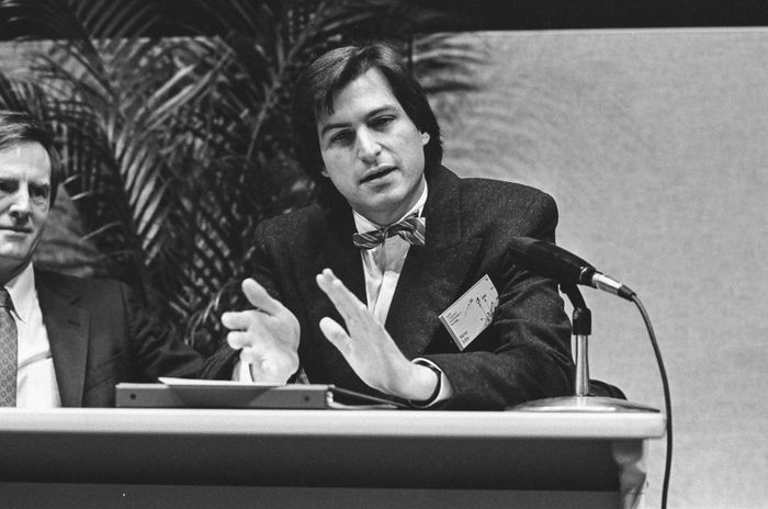 Steve Jobs, dressed in a suit and bow tie, speaks at a panel discussion, gesturing with a microphone in front of him, while another man sits beside him.