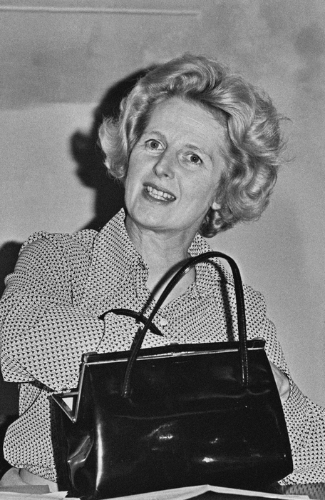 A woman with stylish hair, wearing a polka-dot blouse and holding a handbag, beaming with a smile. Retro chic, possibly attending an event