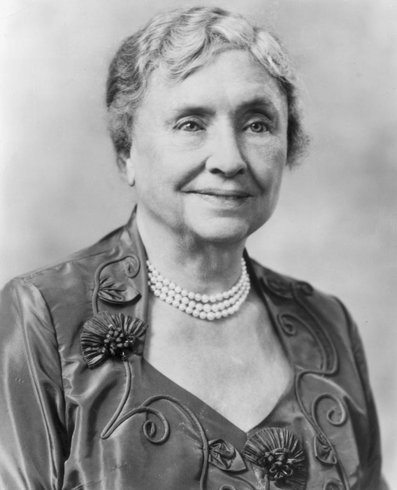An elderly woman with short, wavy hair, wearing a pearl necklace and a dress with floral embroidery, smiling softly in a formal portrait.