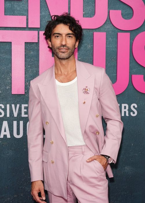 A person dressed in a fashionable pale suit with a mesh shirt and a floral brooch, posing at a media event with a bold text background.
