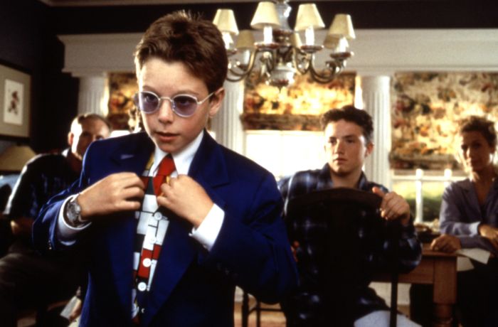 A young child, dressed in a suit and tie, adjusts their attire while others observe in a room