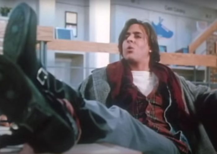 Judd, in a relaxed pose with his feet up, dons a loose-fitting jacket over a plaid shirt and boots, with a school library setting in the background.