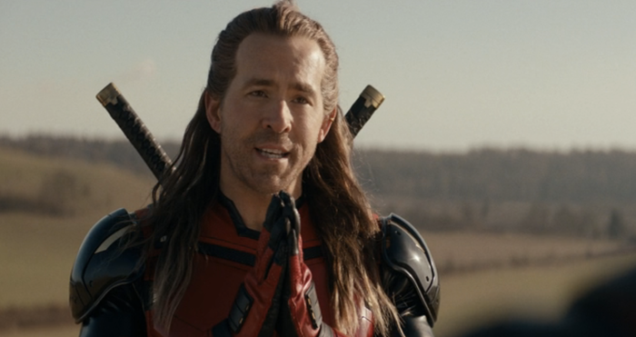 A person in armor with swords on their back talks outdoors in a rural setting.