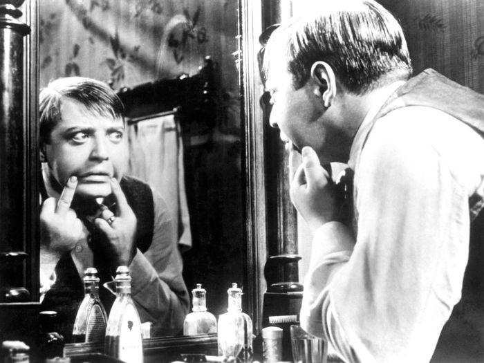 A person is gazing into a mirror, adjusting their facial expression, surrounded by bottles on a vanity table