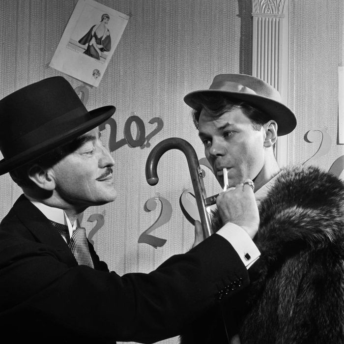Two men dressed in 1940s vintage fashion, one holding a cane and cigarette. They're standing in a room with large numbers and fashion sketches adorning the walls.