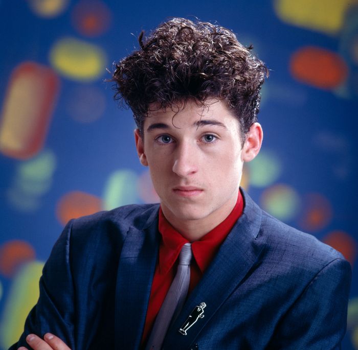 A person with curly hair in a suit and tie, staring at the camera, set against a soft, abstract background