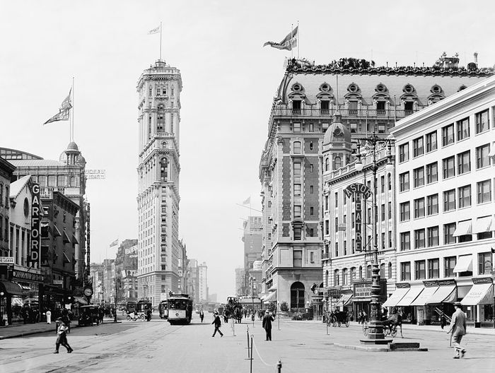 A historic street scene from the early 20th century, featuring vintage architecture, a tall clock tower, streetcars of the time, and pedestrians dressed in period clothing.