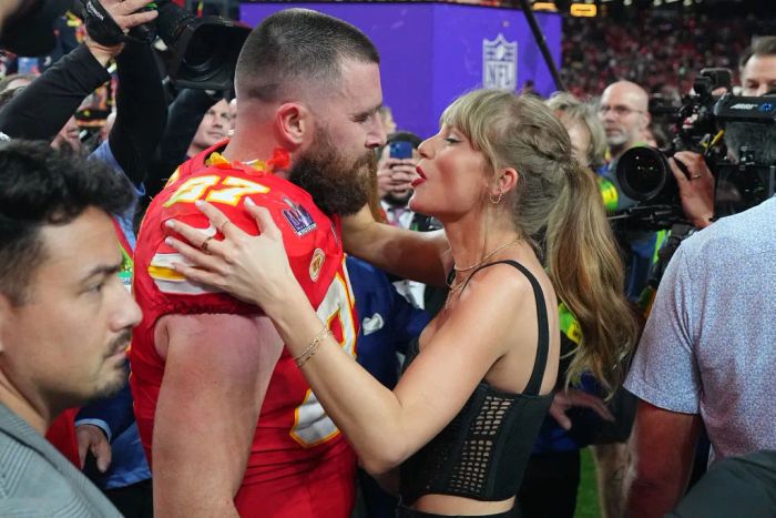 Travis, in uniform, and Taylor, in a chic tank top, share a warm embrace amidst a crowd on the field.