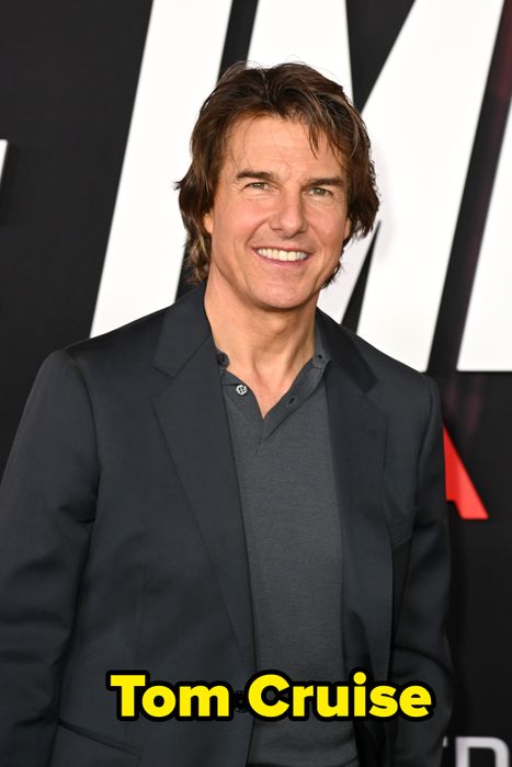 A smiling man in a dark suit and shirt at a formal event with a logo-branded backdrop