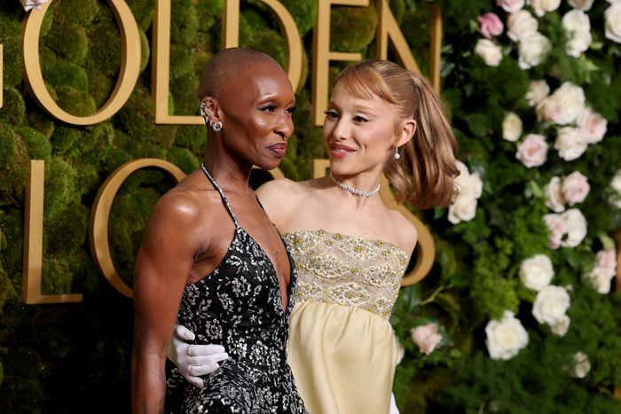 Ariana Grande and Cynthia Erivo pose together on the red carpet, smiling in their elegant attire against a floral backdrop at an event.
