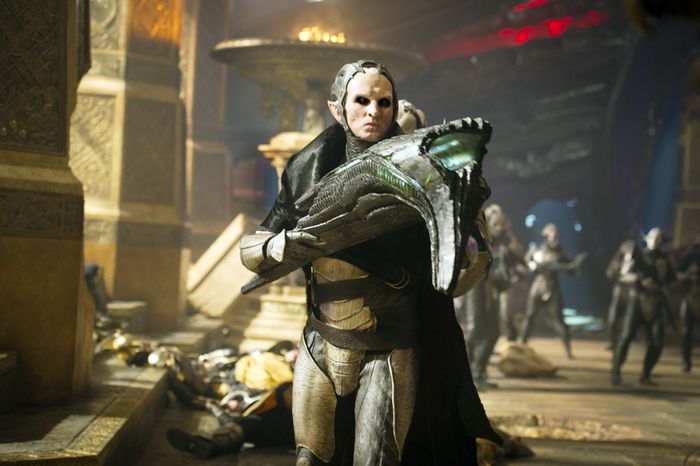 Christopher, clad in armor and a mask, grips a large weapon in a sci-fi environment, with others dressed similarly in the background.