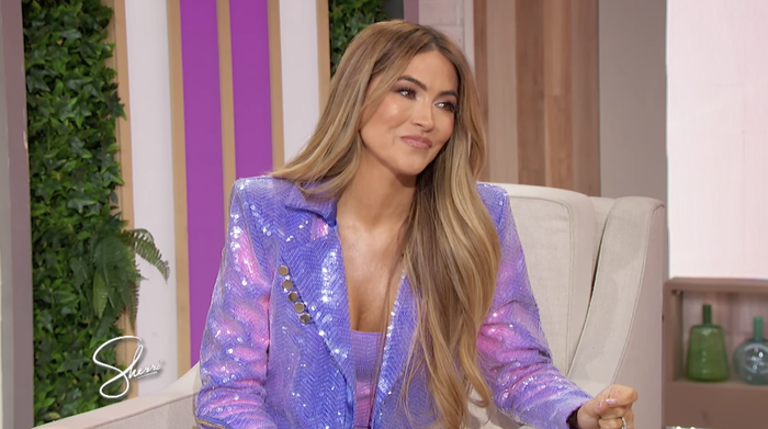 A person seated in an interview setting wearing a sparkling blazer, with long hair styled down, sitting in a brightly lit studio with plants in the background.