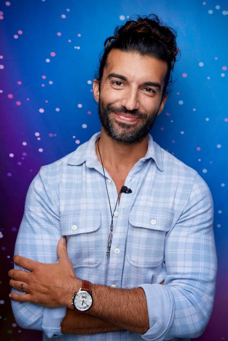 A person with a beard and long hair in a bun, wearing a casual button-up shirt, smiling against a starry backdrop.