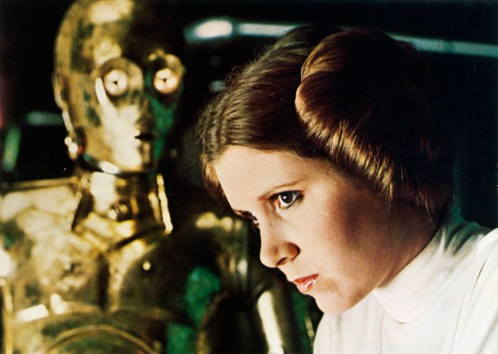 A person with braided hair and a white outfit stands beside a golden robot, both gazing thoughtfully