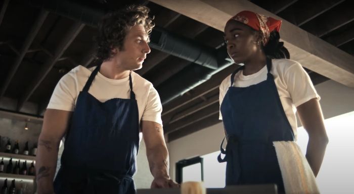 Jeremy and Ayo, both in aprons, engaged in a serious conversation in a kitchen setting.