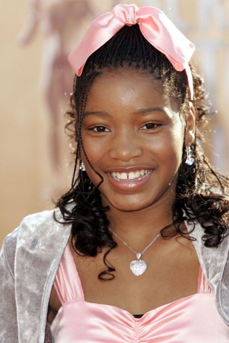 Keke as a teenager, beaming on the red carpet, dressed in a satin gown with a matching hair ribbon, earrings, and a necklace.