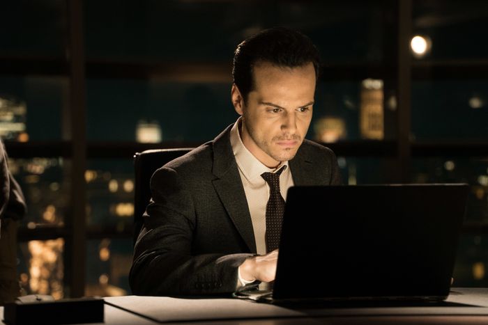 Andrew, dressed in a suit, sits at a desk, deeply focused on a laptop with a city skyline visible through the window.