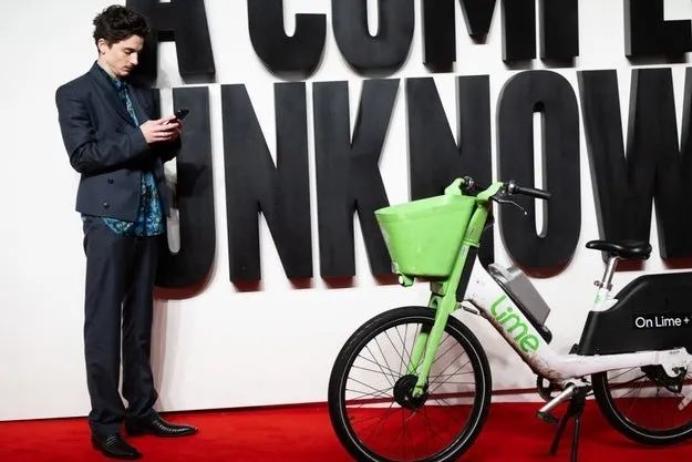 Timothée Chalamet, dressed in a custom suit, stands next to a Lime bike, texting on his phone, in front of a red carpet with large text partially visible in the backdrop