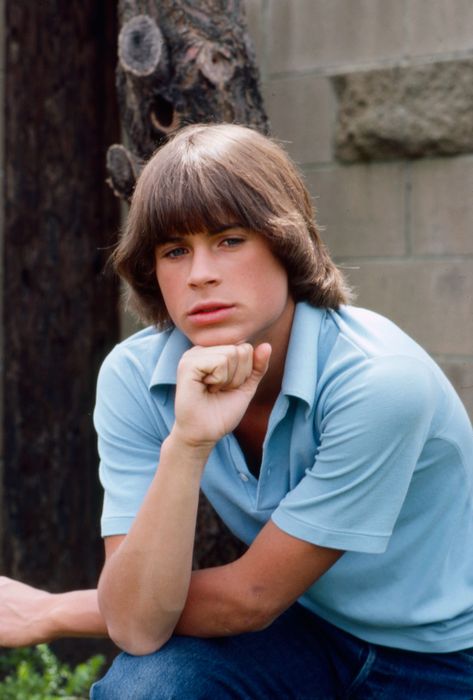 A person with medium-length hair, wearing a casual collared shirt, striking a thoughtful pose with their hand on their chin in an outdoor setting
