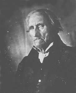 A portrait of an elderly man wearing a suit with a high collar. The photo exudes a vintage, historical atmosphere.