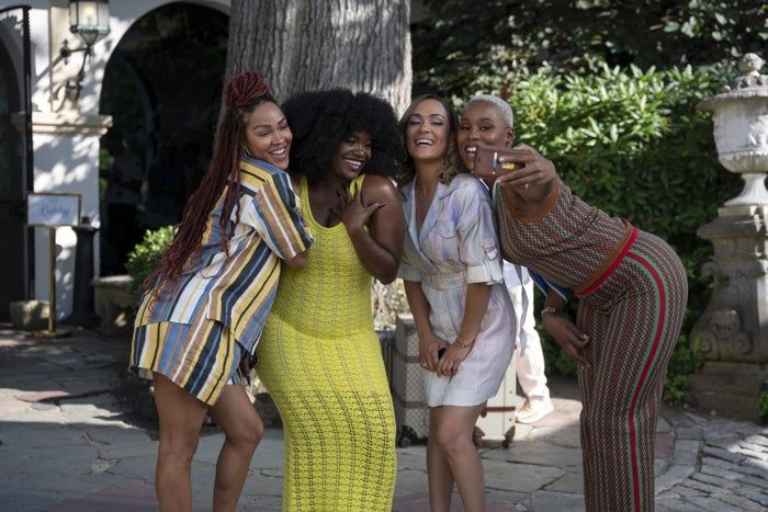 A group of four friends takes a cheerful selfie outdoors, wearing fashionable, patterned outfits. They're all smiling and enjoying a lighthearted moment together.