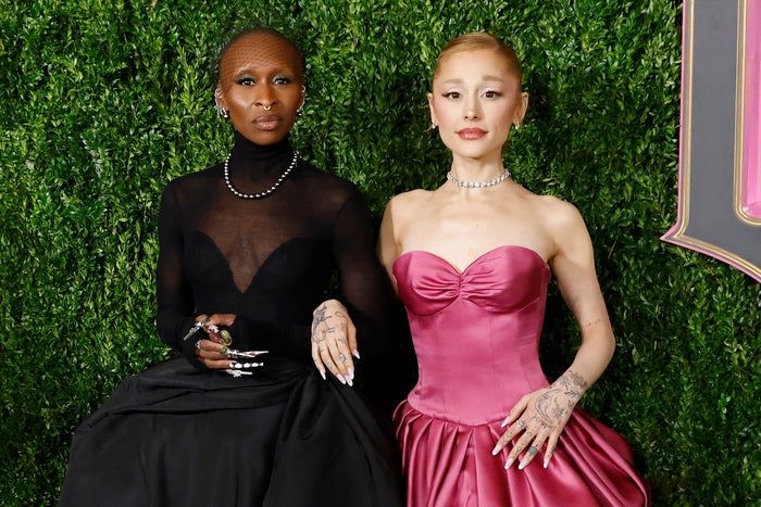 Ariana Grande and Cynthia Erivo, both dressed in beautiful gowns, stand against a vibrant green background: Cynthia in a black outfit with a sheer top, Ariana in a strapless pink dress, both adorned in jewelry