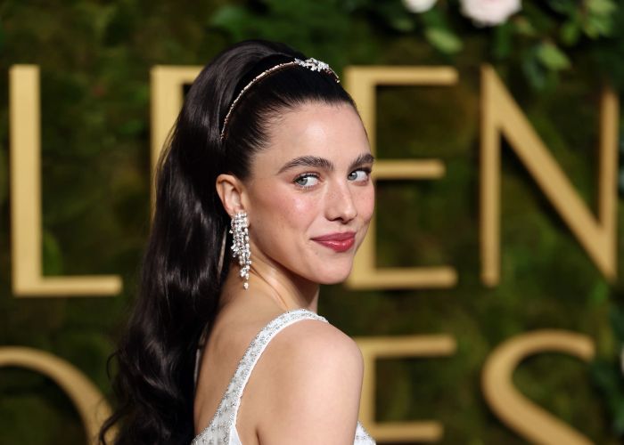 Margaret, with long dark hair and jeweled earrings, strikes a pose on the red carpet in an elegant dress, glancing over her shoulder.