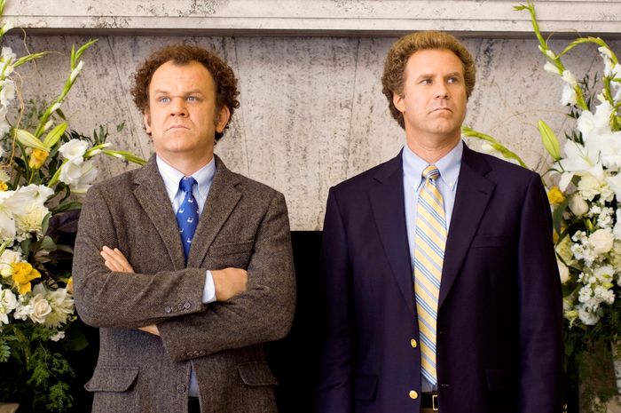 John and Will, both in suits, stand indoors, with John's arms crossed. The background includes floral arrangements.