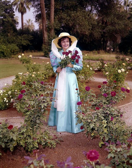A person wearing a vintage-style dress and a large white hat stands in a rose garden, holding a bouquet of flowers, surrounded by lush greenery.