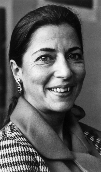 A woman smiling at the camera, wearing a checkered jacket and earrings. Her hair is neatly tied back. A classic black and white photograph