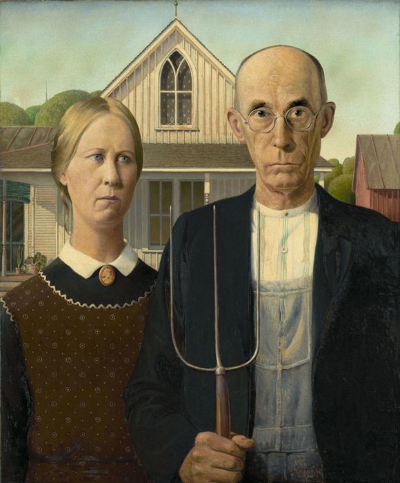 A man and a woman pose in front of a rustic wooden house. The man, holding a pitchfork, is dressed in overalls, while the woman wears a dress with a brooch.