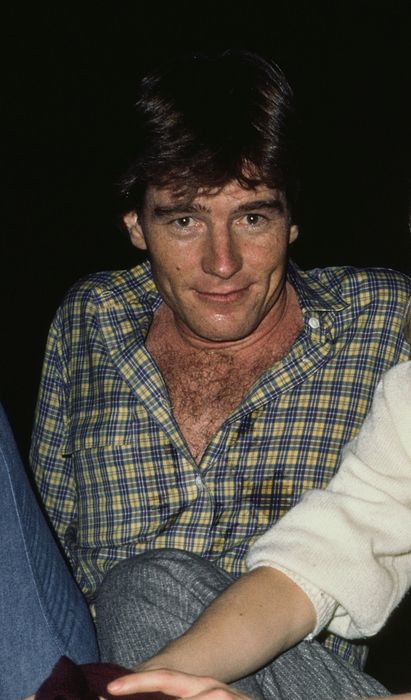Two people seated together. The one on the left is wearing a plaid shirt, while the one on the right is dressed in a light sweater and gray pants