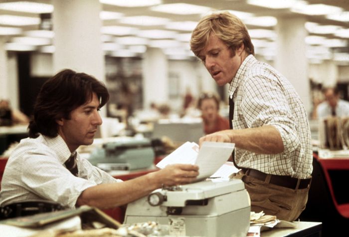 Two men are at a newsroom desk, one seated typing on a typewriter, the other standing, reviewing papers. They are dressed casually, yet in a businesslike manner