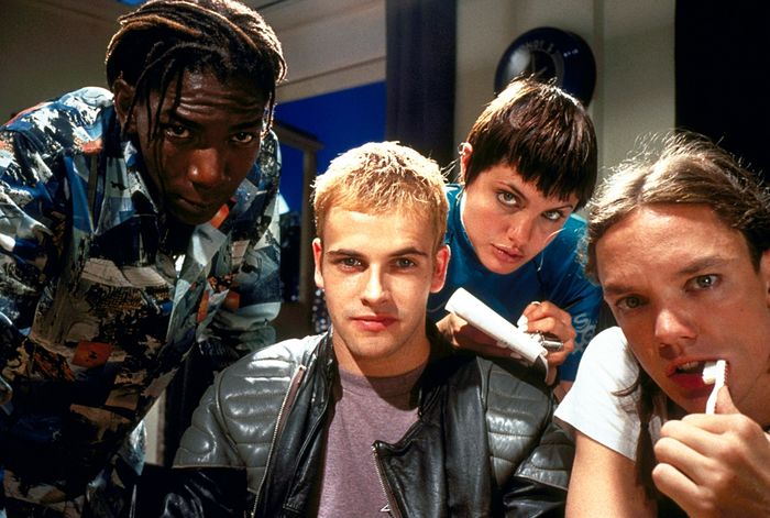 A group of four individuals dressed in fashionable 90s clothing, including leather jackets and patterned outfits, striking a pose with a cool, edgy vibe
