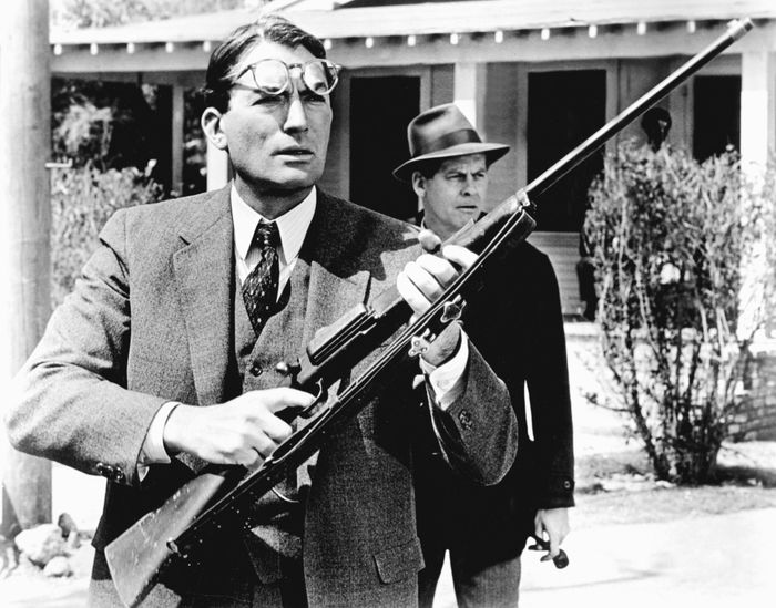 A man dressed in a suit, holding a rifle, with another man in a hat standing behind him outdoors