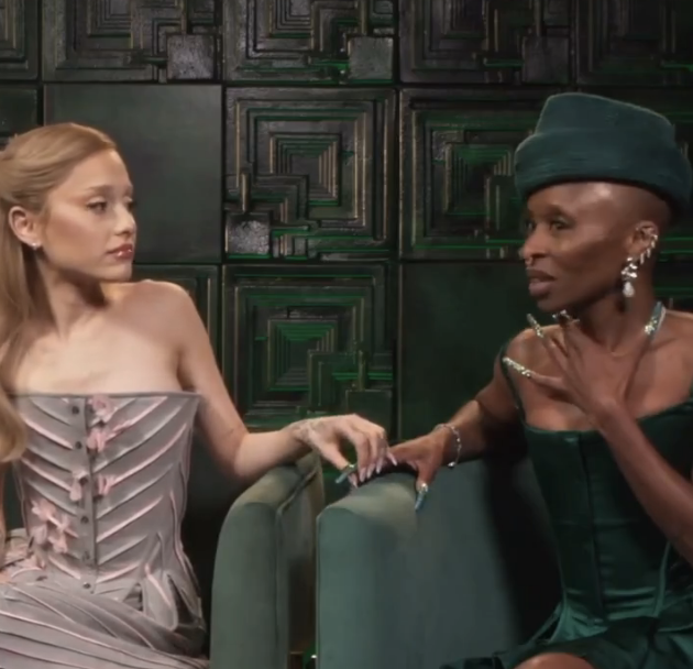 Ariana Grande and Cynthia Erivo are seated, deep in conversation. Ariana wears a strapless, tailored dress while Cynthia dons a strapless gown with a coordinating headpiece. Ariana holds Cynthia's index finger.