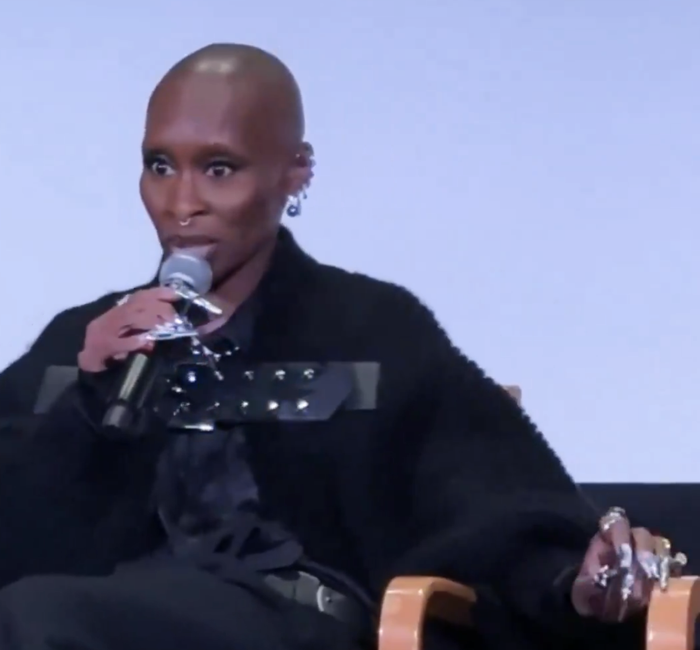 Cynthia Erivo speaks into a microphone while seated on stage, sporting oversized rings and a fashionable jacket.