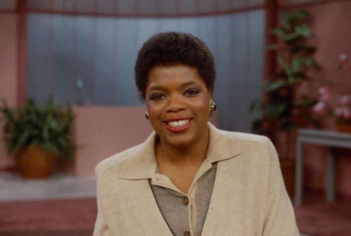 Oprah Winfrey smiles warmly in a studio, wearing a beige blazer over a grey top, with plants and curtains in the background.