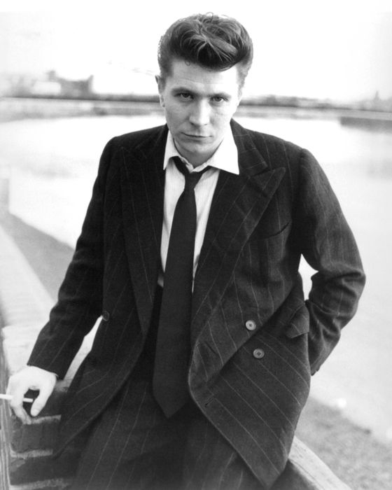An individual in a pinstripe suit and tie, casually leaning against a brick wall with a cigarette in hand. Looks like it could be from a classic setting by the water