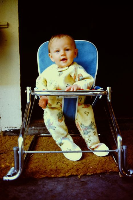A baby sitting in a high chair, dressed in a patterned onesie, looking towards the camera indoors
