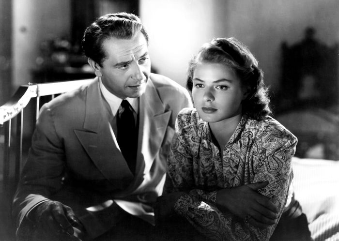Two people, dressed in 1940s-style outfits, sitting together indoors, intently gazing at something out of the frame