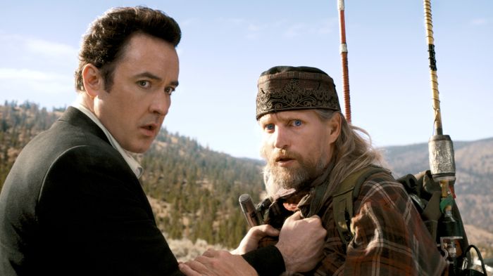 Two individuals in a high-stakes outdoor setting, one dressed in a suit, the other in a plaid shirt and bandana, both holding hiking equipment