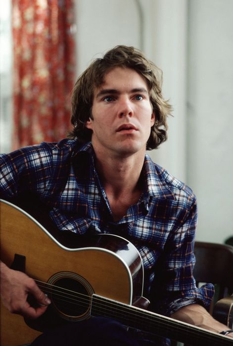 A person wearing a plaid shirt strums an acoustic guitar while gazing thoughtfully upwards, indoors with a curtain behind them