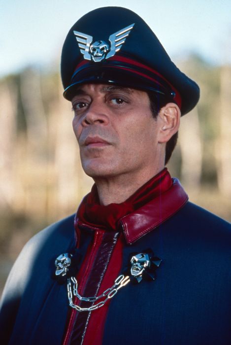 Raul, dressed as a military figure, wears a cap adorned with wings and a skull emblem, along with a chain of skulls.