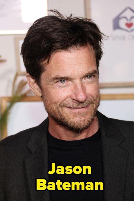 A person with a short hairstyle and facial stubble, wearing a black shirt and dark jacket, smiles faintly