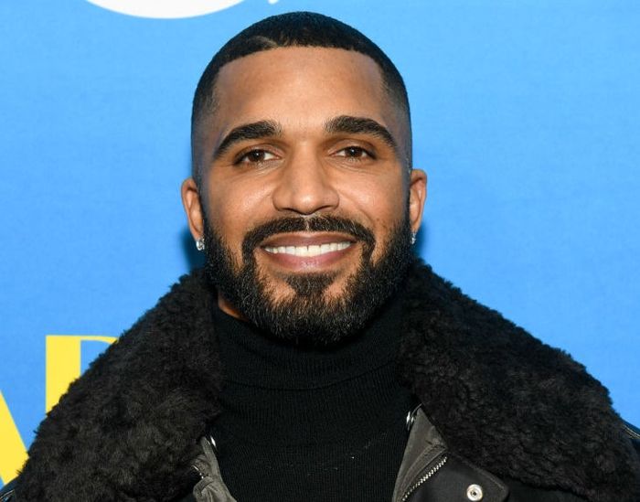 A man with a beard, dressed in a trendy leather jacket with a shearling collar, smiling at a public event.
